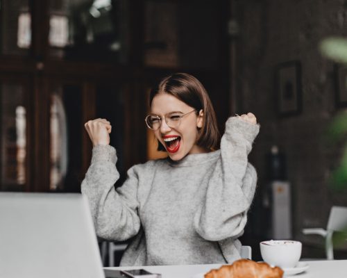 Happy woman in glasses makes winning gesture and sincerely rejoices. Lady with red lipstick dressed in gray sweater looking at laptop